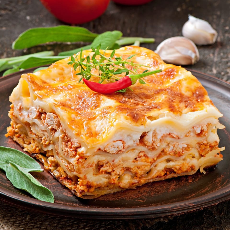 A golden-brown slice of Caruso's Italian lasagna, garnished with a red chili and thyme.