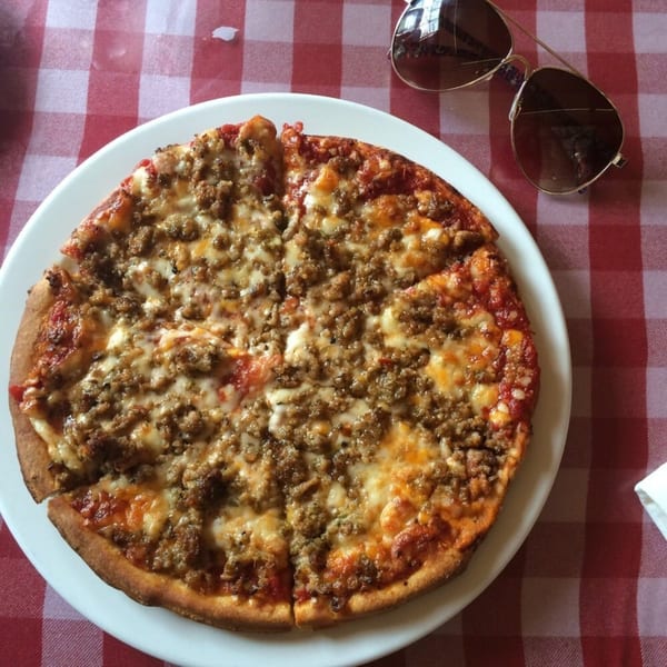A Caruso's Italian sausage pizza on a white plate, with sunglasses on a red and white checkered tablecloth.