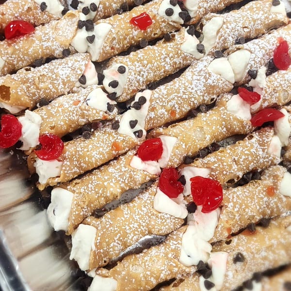 Rows of Caruso's cannolis, filled with cream, topped with chocolate chips, powdered sugar, and cherries.